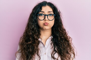 Young brunette woman with curly hair wearing casual clothes and glasses puffing cheeks with funny...