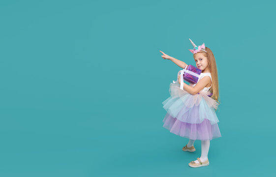 Cute child girl in elegant tulle dress and unicorn headband celebrates birthday  party on colored background. Funny face with gift box.