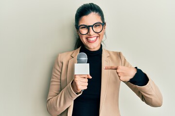 Young hispanic woman holding reporter microphone pointing finger to one self smiling happy and proud