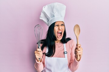 Young hispanic woman wearing baker uniform holding spoon and whisk angry and mad screaming...