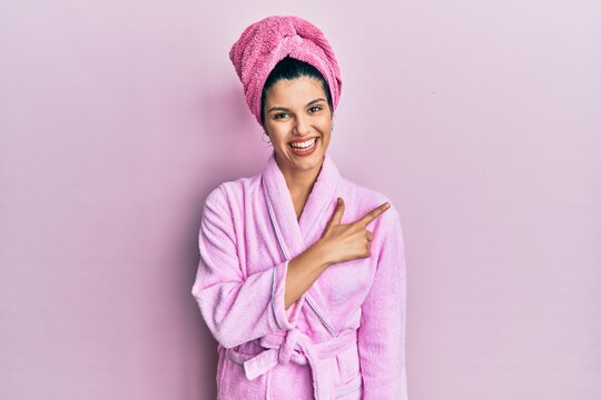Young hispanic woman wearing shower towel cap and bathrobe cheerful with a smile of face pointing with hand and finger up to the side with happy and natural expression on face