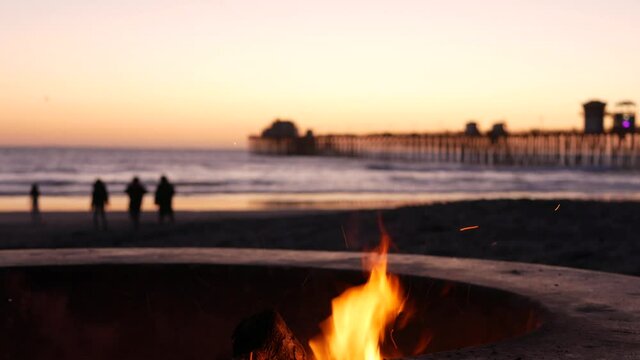 Campfire pit by Oceanside pier, California USA. Camp fire burning on ocean beach, bonfire flame in cement ring place for bbq, sea water waves. Romantic evening twilight sky, dusk after summer sunset.