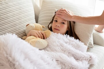 A sick girl is lying under a blanket holding a teddy bear. Checking fever with mom's hand.