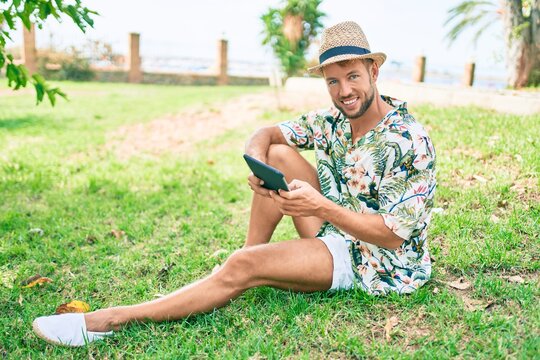Caucasian handsome man smiling happy using touchpad device sitting on the grass at the park