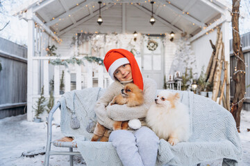 Joyful child, a cute boy dressed in a red santa claus hat holds two Spitz puppies in his hands on the background of a white house in winter. Christmas gift for a child - 402911417