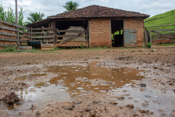 A kind of barn on a corral, after a heavy rain with the ground full of puddles. In the background there are a green pasture and blue sky