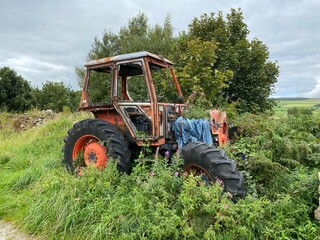 Obraz premium An old discarded orange tractor, overgrown with long grasses, with hills and meadows, in the distance near, Silsden, Keighley, UK