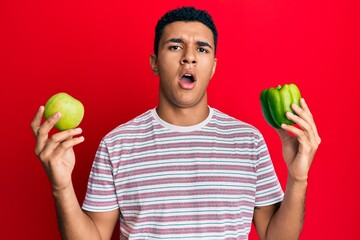 Young arab man holding green apple and pepper in shock face, looking skeptical and sarcastic, surprised with open mouth