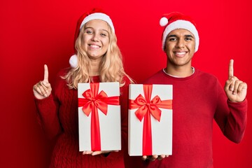Young interracial couple wearing christmas hat and holding a gift smiling with an idea or question pointing finger with happy face, number one