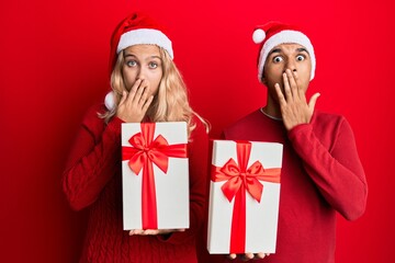 Young interracial couple wearing christmas hat and holding a gift covering mouth with hand, shocked and afraid for mistake. surprised expression