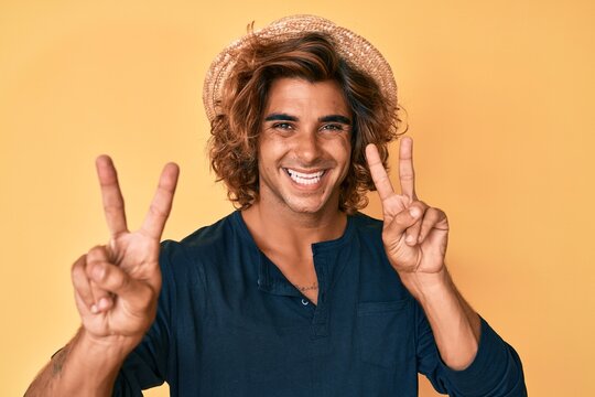 Young hispanic man wearing summer hat smiling looking to the camera showing fingers doing victory sign. number two.