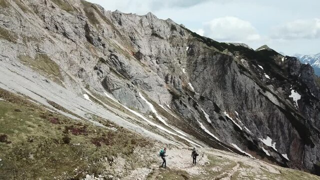 A panoramic drone shot of a couple hiking in the region of Sinabell, Austrian Alps. The slopes are still partially covered with snow. Stony and sharp mountains. A bit of overcast. Exploring the nature