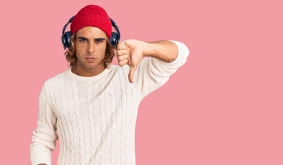 Young hispanic man listening to music using headphones looking unhappy and angry showing rejection and negative with thumbs down gesture. bad expression.