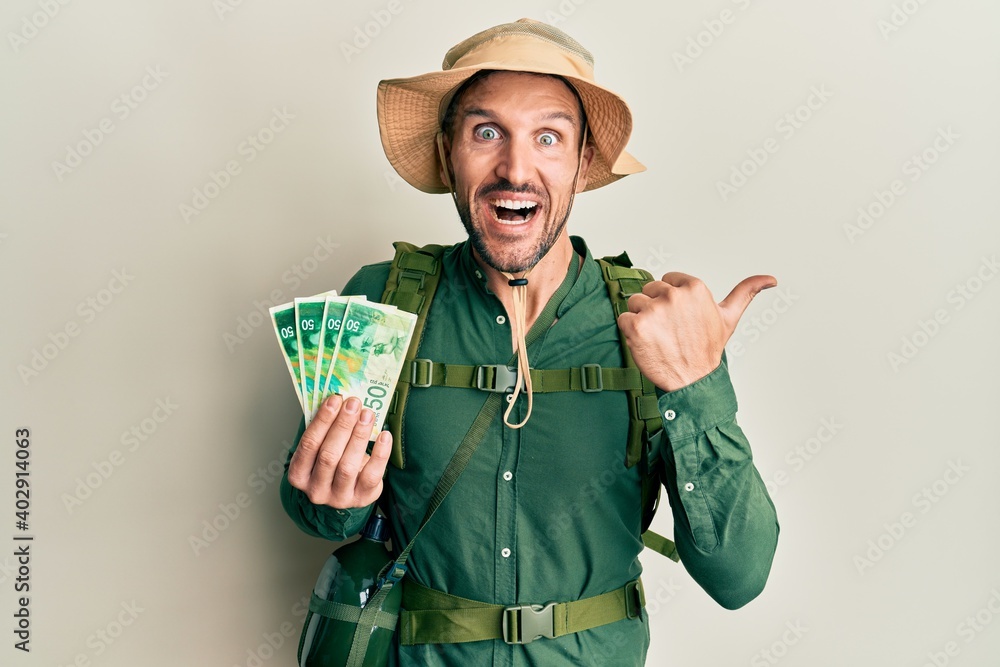 Sticker Handsome man with beard wearing explorer hat holding 50 shekels pointing thumb up to the side smiling happy with open mouth