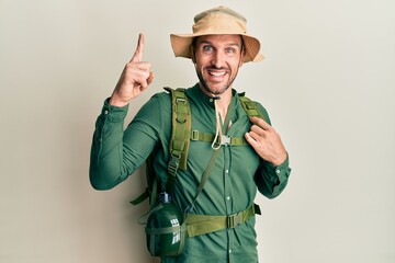 Handsome man with beard wearing explorer hat and backpack smiling amazed and surprised and pointing up with fingers and raised arms.