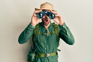 Handsome man with beard wearing explorer hat looking through binoculars in shock face, looking skeptical and sarcastic, surprised with open mouth