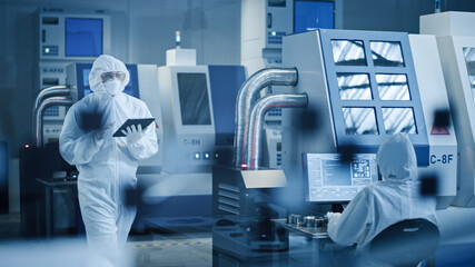 Research Factory Cleanroom: Engineer Wearing Coverall and Mask, Walking Through Workshopop Uses Tablet Computer. In Background Professionals Work on Modern High Tech CNC Machinery