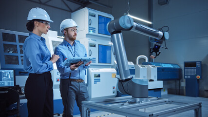 Factory Workshop: Professional Female Engineer, Male Machinery Operator Use Industrial Digital Tablet Computer to Work and Program Robot Arm for Production Line. High-Tech Facility with CNC Machines