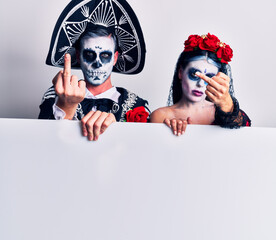 Young couple wearing mexican day of the dead costume holding blank empty banner showing middle finger, impolite and rude fuck off expression