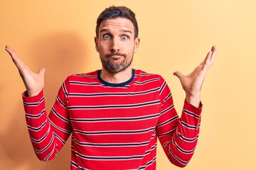 Young handsome man wearing casual striped t-shirt standing over isolated yellow background clueless and confused with open arms, no idea and doubtful face.