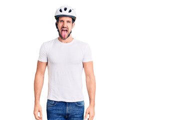 Young handsome man wearing bike helmet sticking tongue out happy with funny expression. emotion concept.