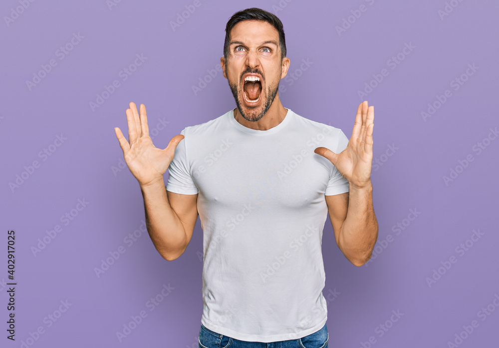 Poster Handsome man with beard wearing casual white t shirt crazy and mad shouting and yelling with aggressive expression and arms raised. frustration concept.