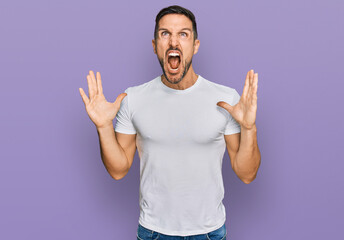 Handsome man with beard wearing casual white t shirt crazy and mad shouting and yelling with...