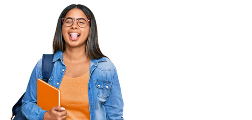 Young latin girl wearing student backpack and holding books sticking tongue out happy with funny expression. emotion concept.