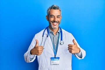 Middle age grey-haired man wearing doctor uniform and stethoscope success sign doing positive gesture with hand, thumbs up smiling and happy. cheerful expression and winner gesture.