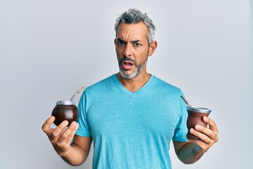 Middle age grey-haired man drinking mate infusion in shock face, looking skeptical and sarcastic,...