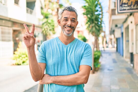Middle age grey-haired man wearing casual clothes at street of city smiling with happy face winking at the camera doing victory sign. number two.