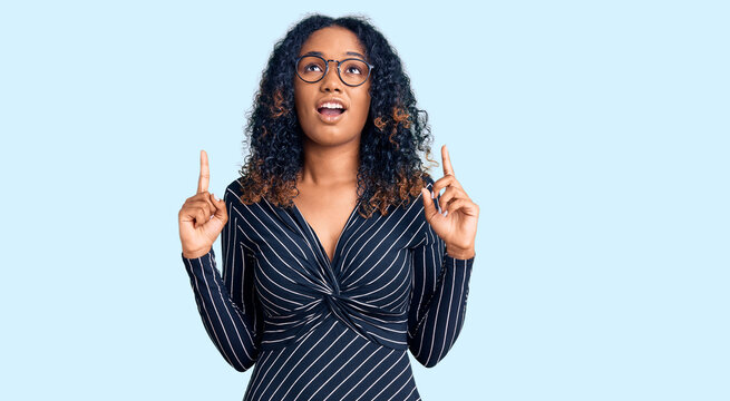 Young african american woman wearing casual clothes and glasses amazed and surprised looking up and pointing with fingers and raised arms.