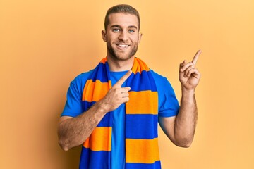 Young caucasian man football hooligan cheering game smiling and looking at the camera pointing with two hands and fingers to the side.