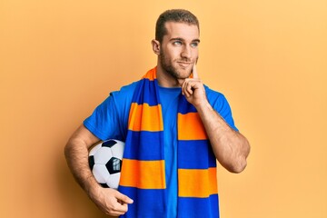 Young caucasian man football hooligan cheering game holding ball serious face thinking about question with hand on chin, thoughtful about confusing idea