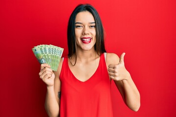 Young hispanic girl holding 1000 chilean pesos smiling happy and positive, thumb up doing excellent and approval sign
