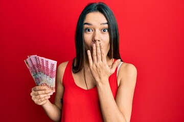 Young hispanic girl holding 100000 indonesian rupiah covering mouth with hand, shocked and afraid...
