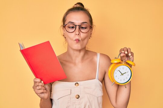 Beautiful caucasian woman holding book and alarm clock making fish face with mouth and squinting eyes, crazy and comical.