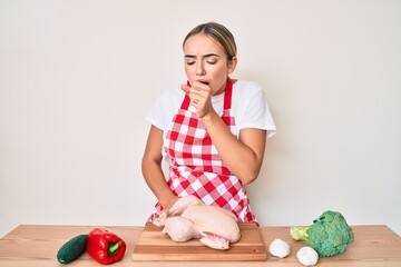 Fototapeta premium Young beautiful blonde woman wearing apron cooking chicken feeling unwell and coughing as symptom for cold or bronchitis. health care concept.