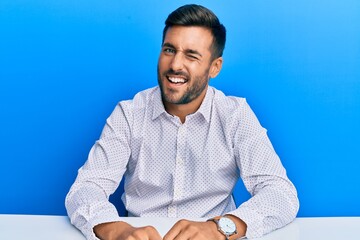 Handsome hispanic man wearing business clothes sitting on the table winking looking at the camera...