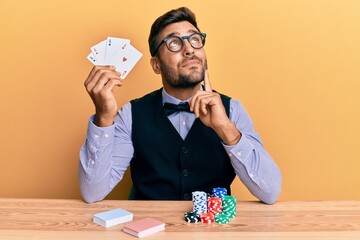 Handsome hispanic croupier man sitting on the table with poker chips and cards serious face...