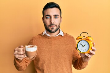 Young hispanic man holding coffee and alarm clock relaxed with serious expression on face. simple...