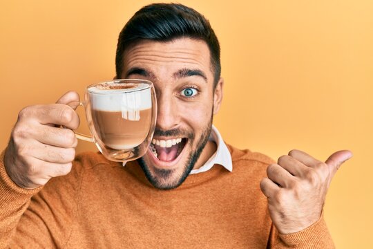 Young hispanic man holding cup of coffee over eye pointing thumb up to the side smiling happy with open mouth