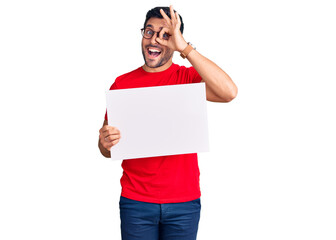 Young hispanic man holding blank empty banner smiling happy doing ok sign with hand on eye looking through fingers