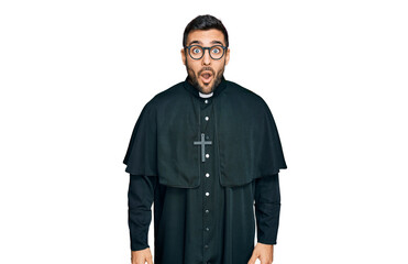 Young hispanic man wearing priest uniform scared and amazed with open mouth for surprise, disbelief face