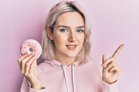 Young blonde girl holding donut smiling happy pointing with hand and finger to the side