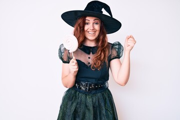 Young beautiful woman wearing witch halloween costume holding lollipop screaming proud, celebrating victory and success very excited with raised arms