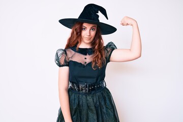 Young beautiful woman wearing witch halloween costume strong person showing arm muscle, confident...