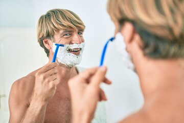 Handsome blond man looking at the mirror at the bathroom saving using foam and razor