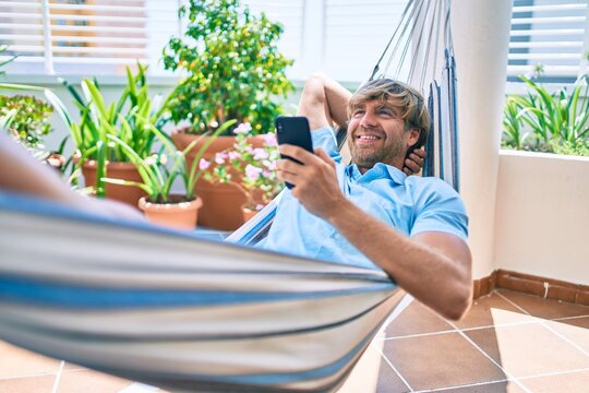 Middle age handsome man at the terrace of his house relaxing lying on a hammock with the phone