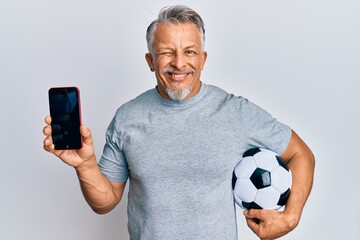 Middle age grey-haired man holding football ball showing smartphone winking looking at the camera...